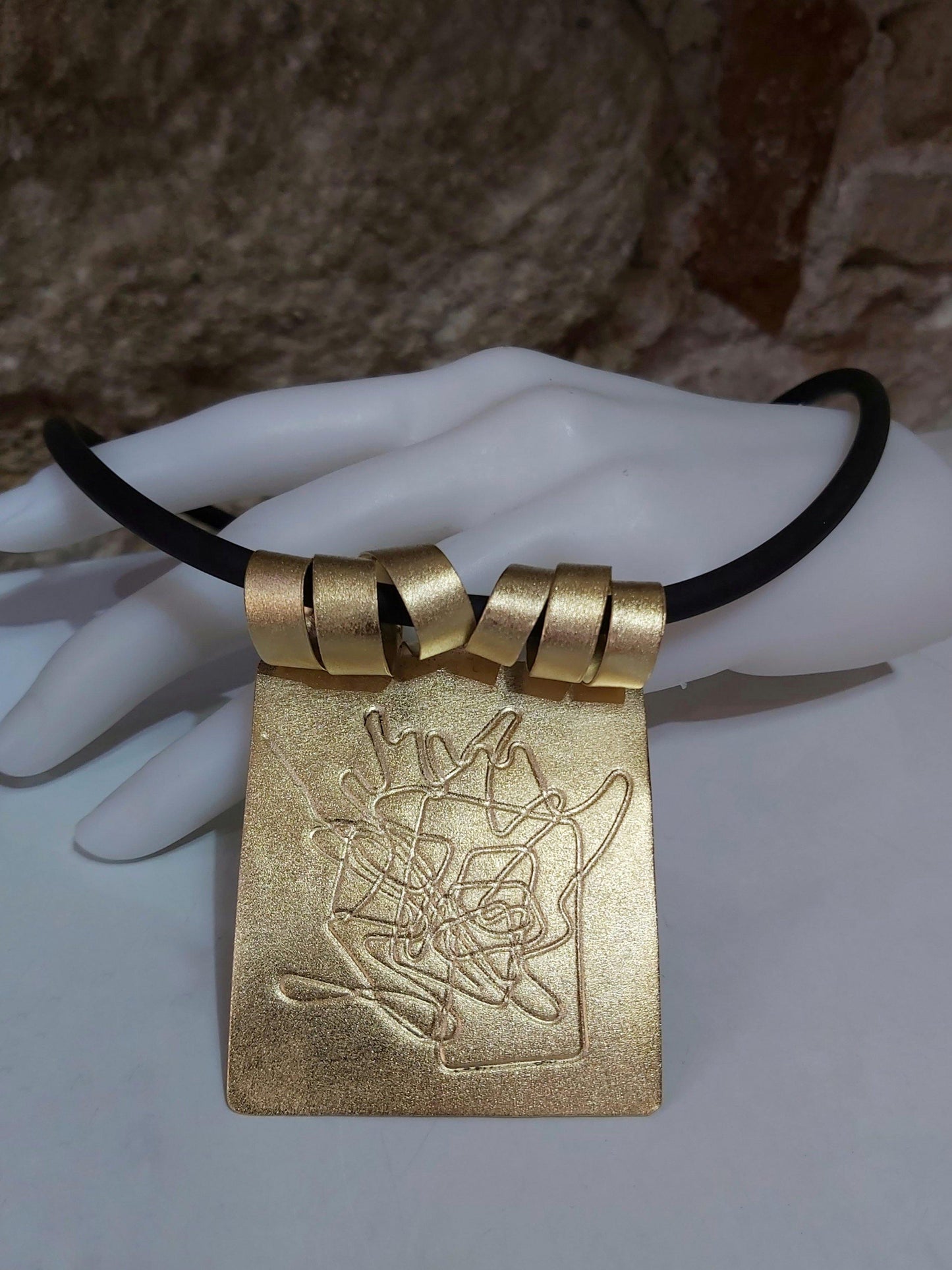 Colgante artesanal chapado en oro con grabado original montado sobre caucho negro. Hecho a mano por Amparo Valencia