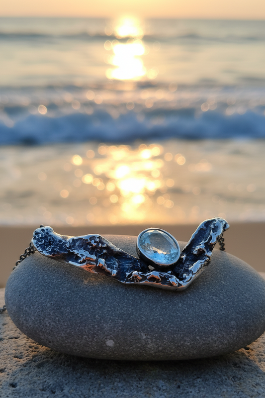 Colgante artesanal de plata 925 con una aguamarina natural montado sobre una piedra lisa, fotografiado con un fondo cálido de atardecer en la playa